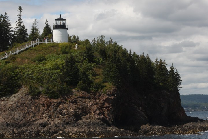 Lighthouse on a rocky, forested cliff under a cloudy sky.