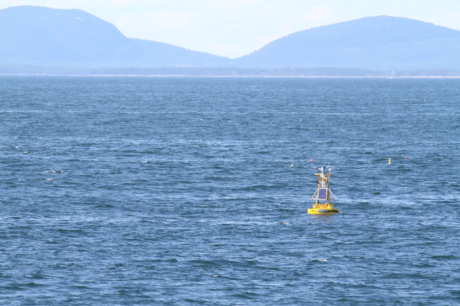 Yellow buoy with solar panel floats on a large blue body of water, mountains in background.